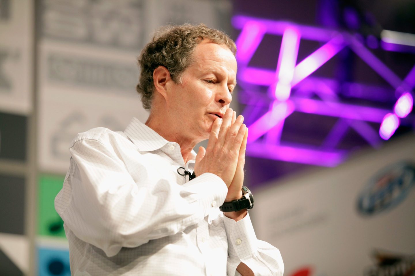 John Mackey, standing on stage, has his head turned downward with both hands touching his lips.