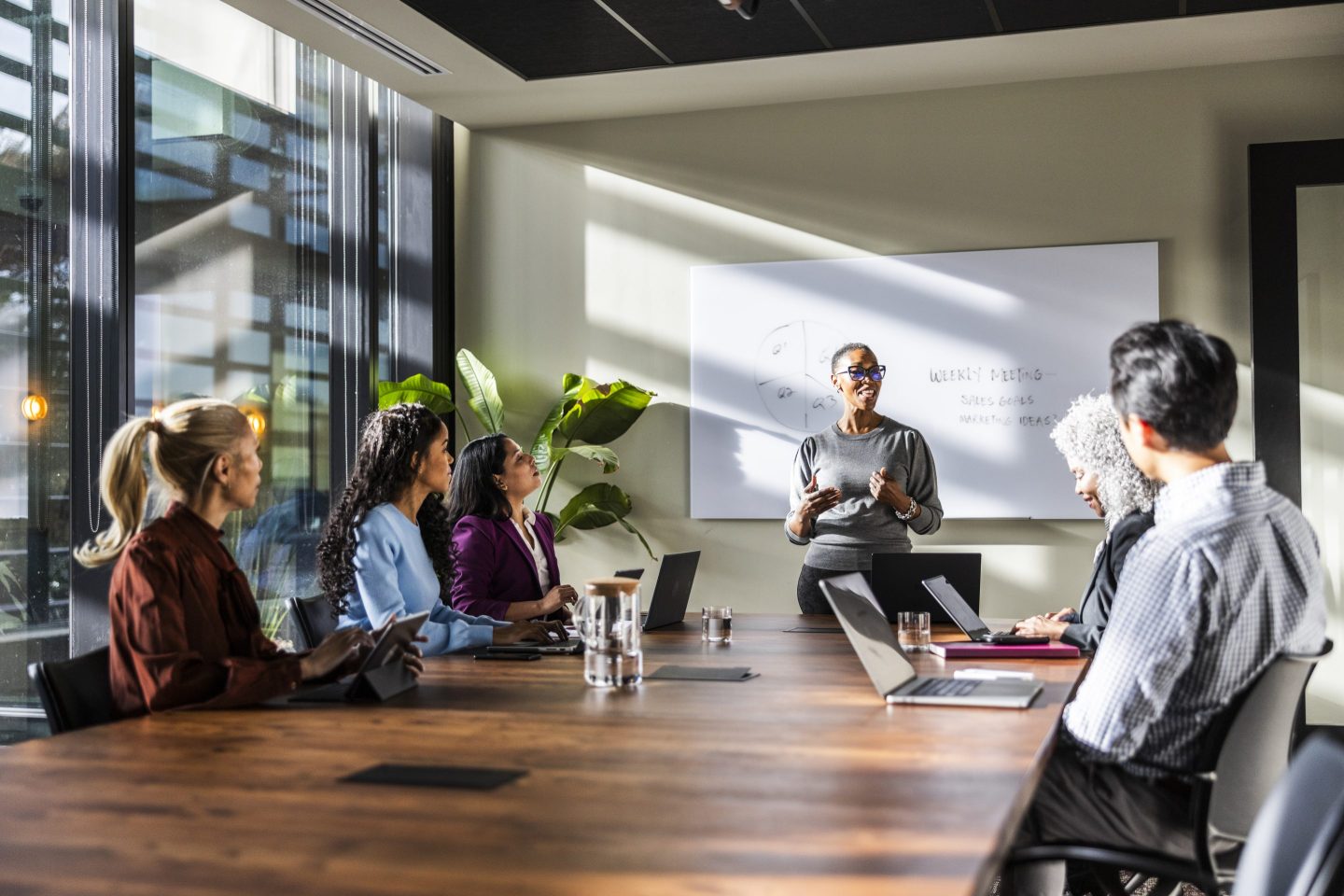 A woman stands in front of a whiteboard speaking to a table of people.