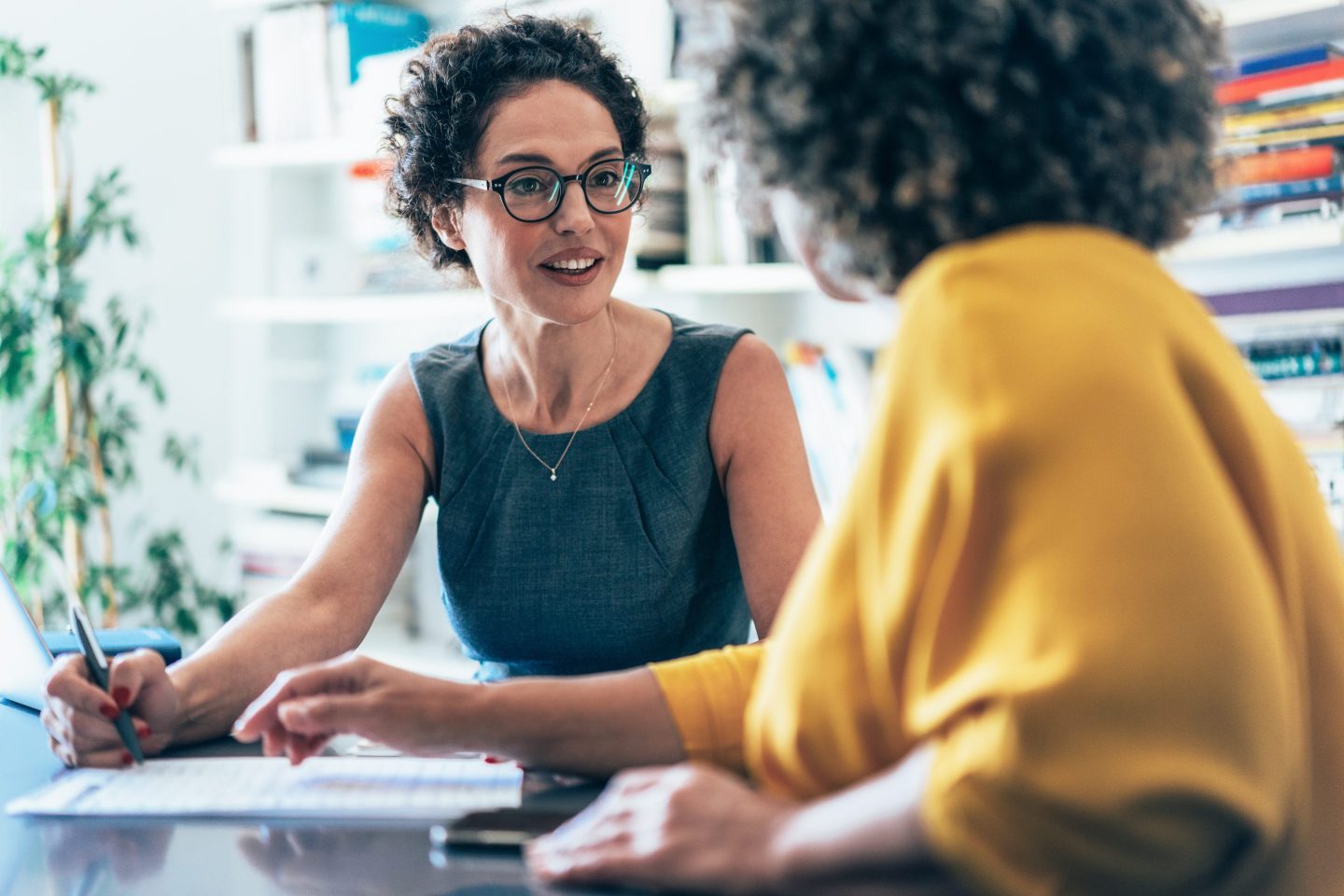 Two women office workers sit and talk at a desk.