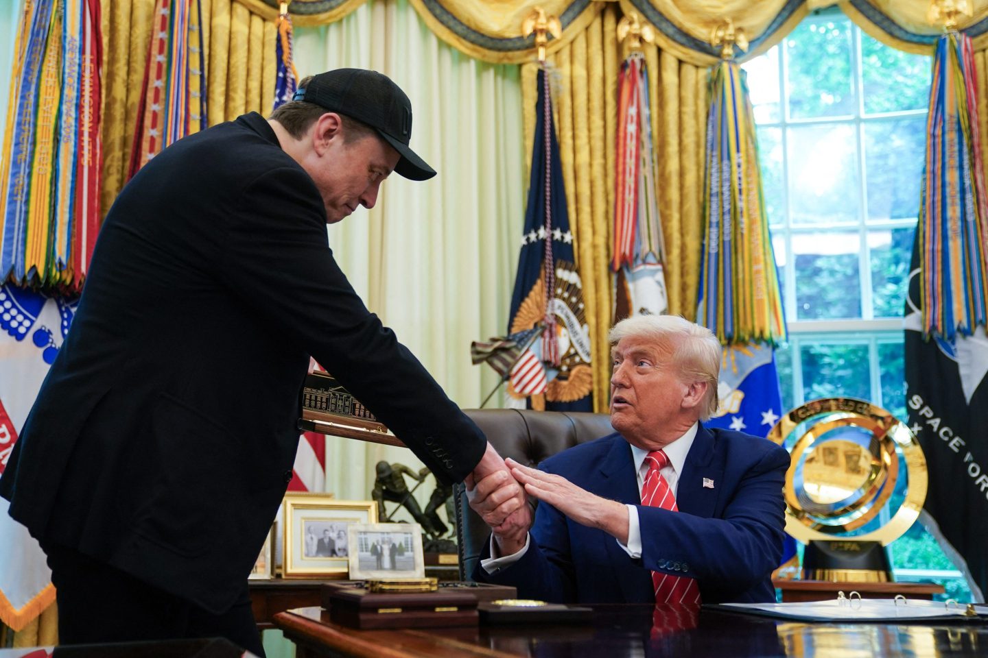 Trump, sitting at his desk in the Oval Office, shakes Elon Musk's hand.