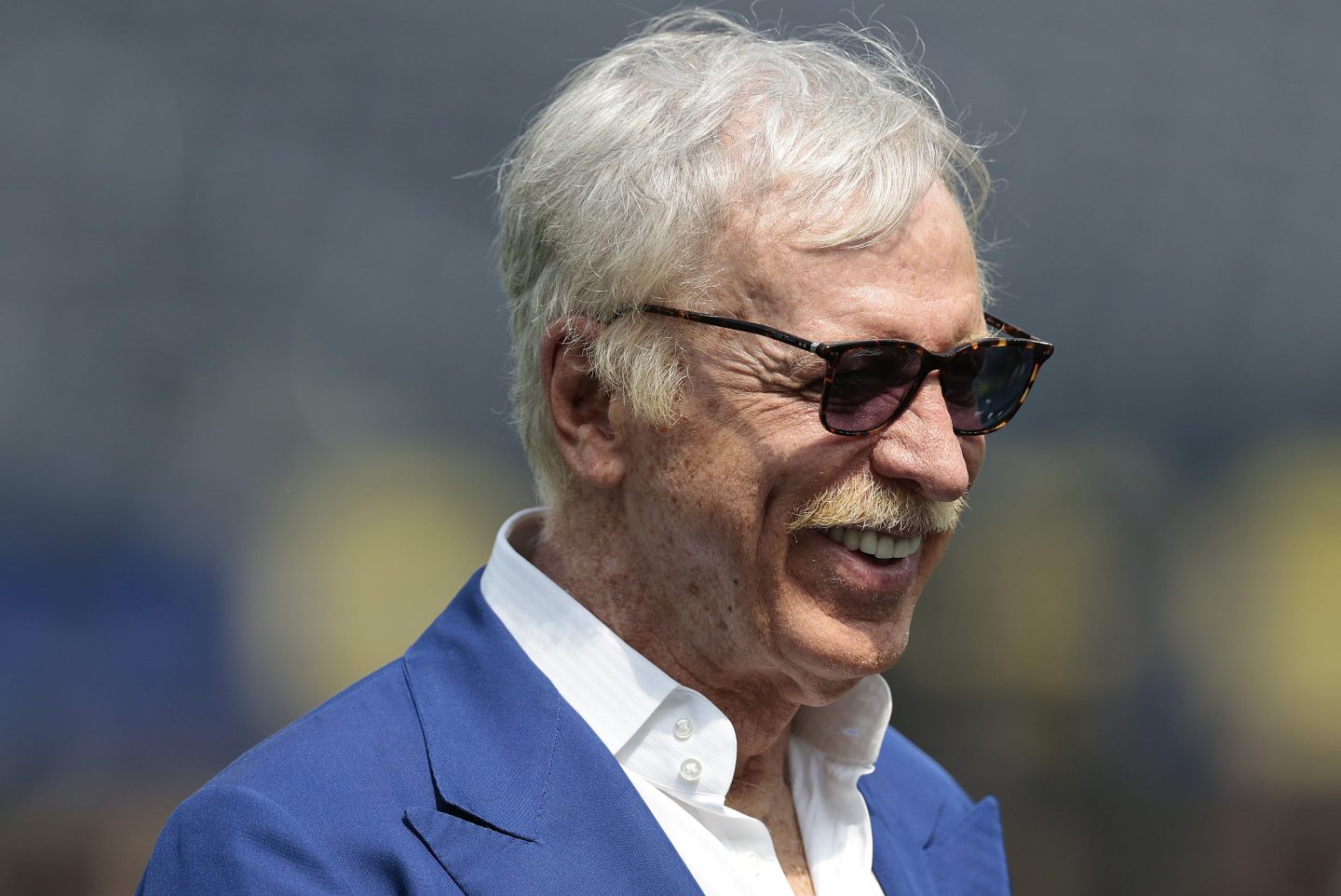 Stan Kroenke, wearing a blue suit and sunglasses, smiles.