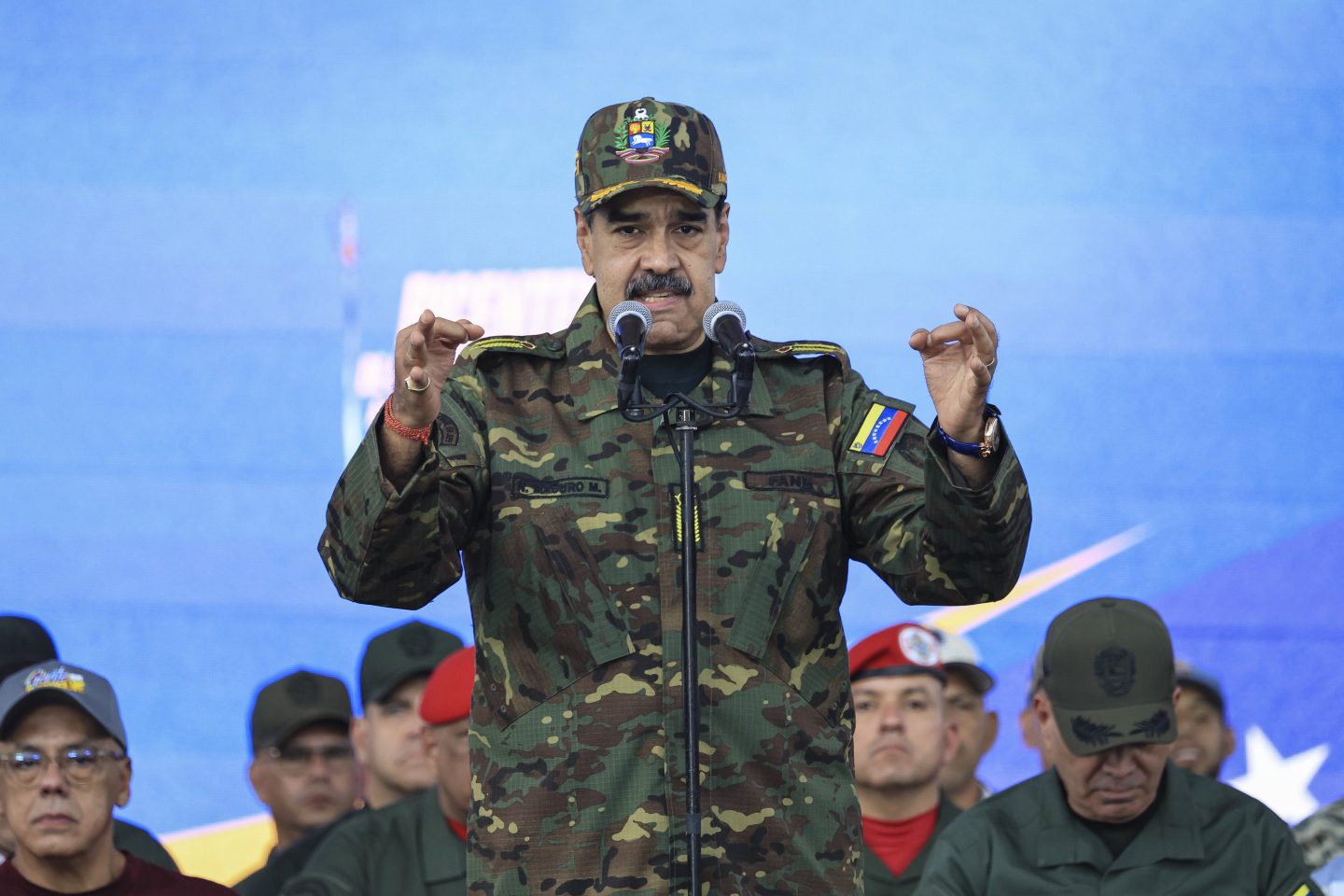 Nicolas Maduro, dressed in military uniform, speaks behind a microphone with both hands raised.