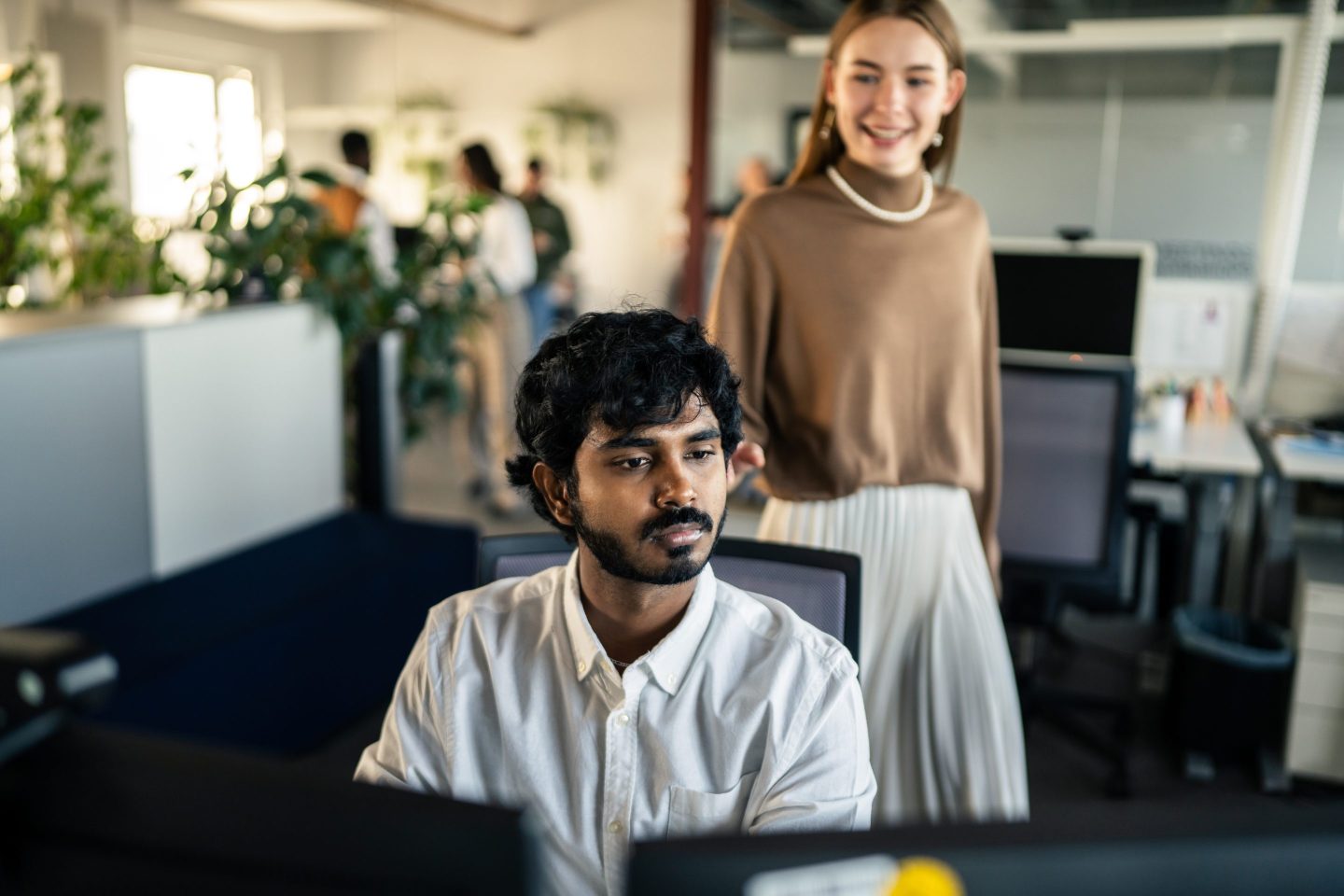 A man works on two computers while a coworker looks on in the background.