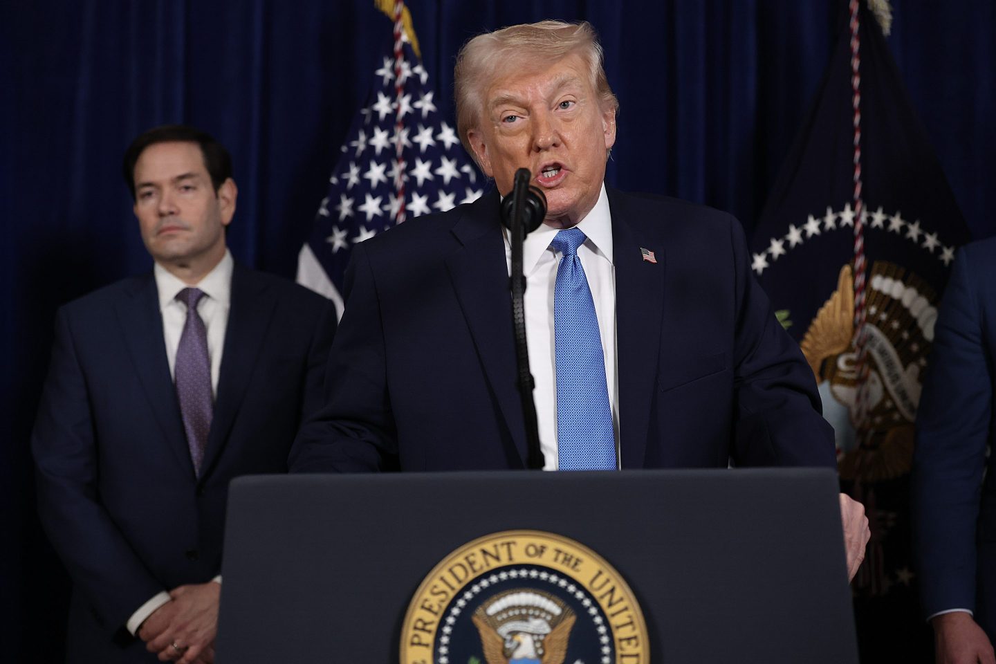 Donald Trump speaks behind a podium with Marco Rubio frowning in the background.