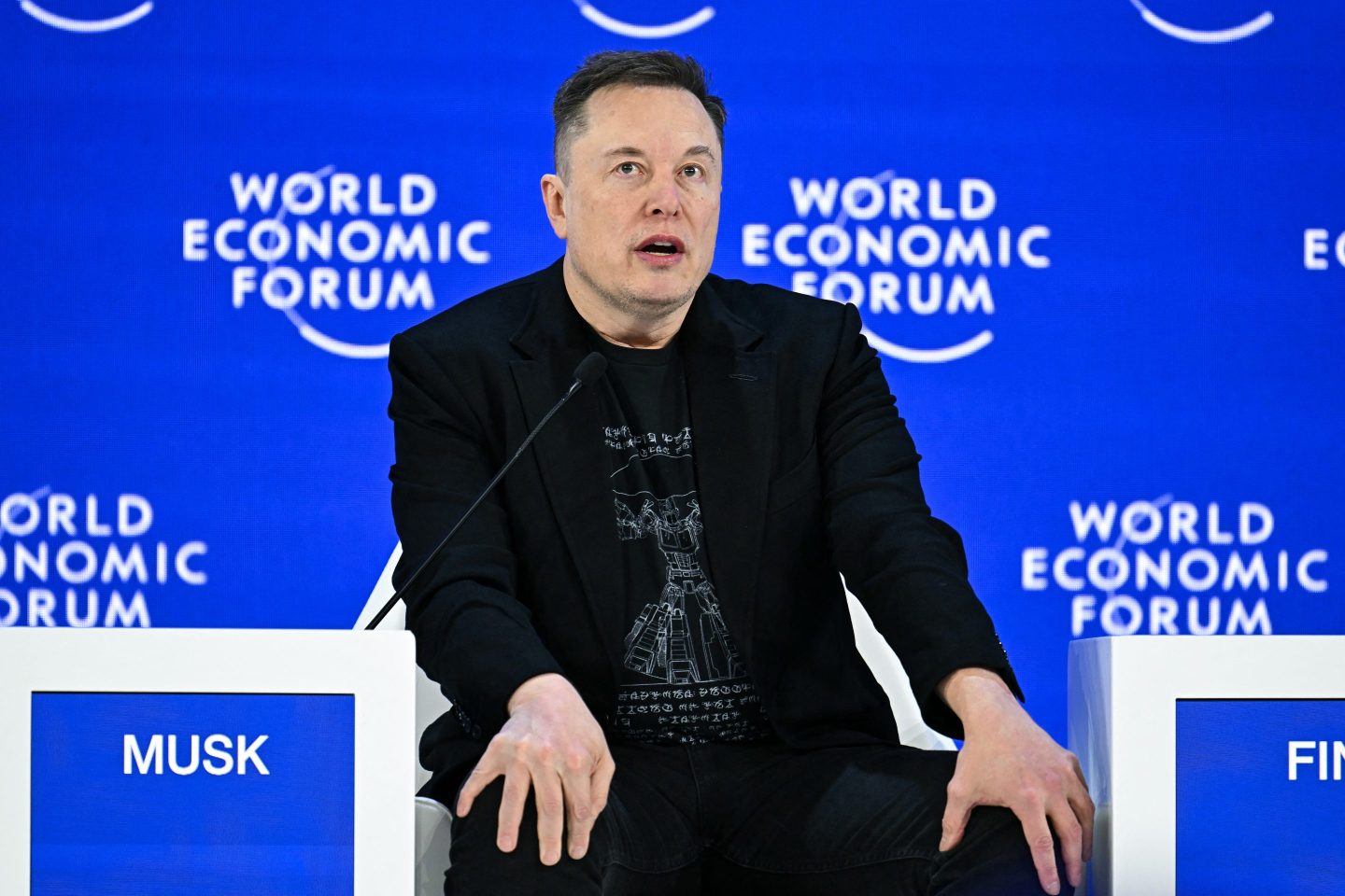 Elon Musk sits with his hands on his knees in front of a blue "World Economic Forum" background.