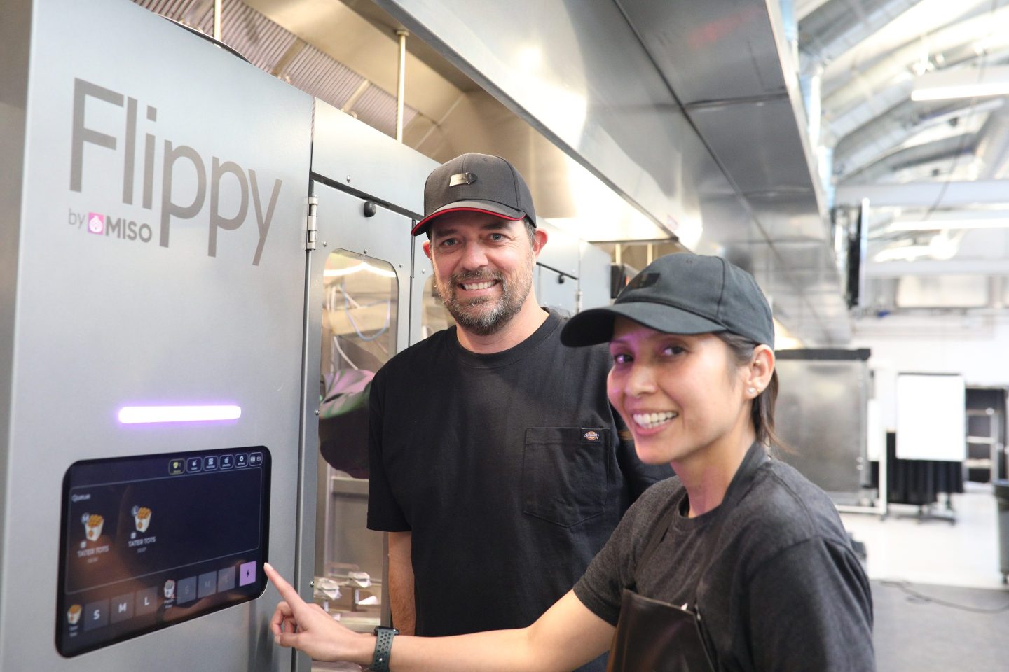 Two restaurant workers wearing black stand in front of a silver "Flippy" fry station.