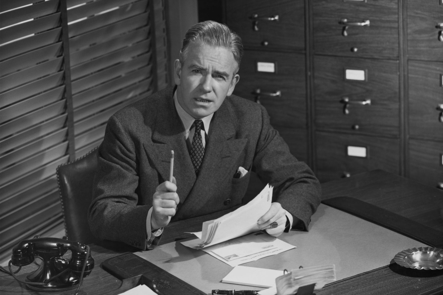 A man in a suit sits behind a desk and waves his pencil towards the camera.