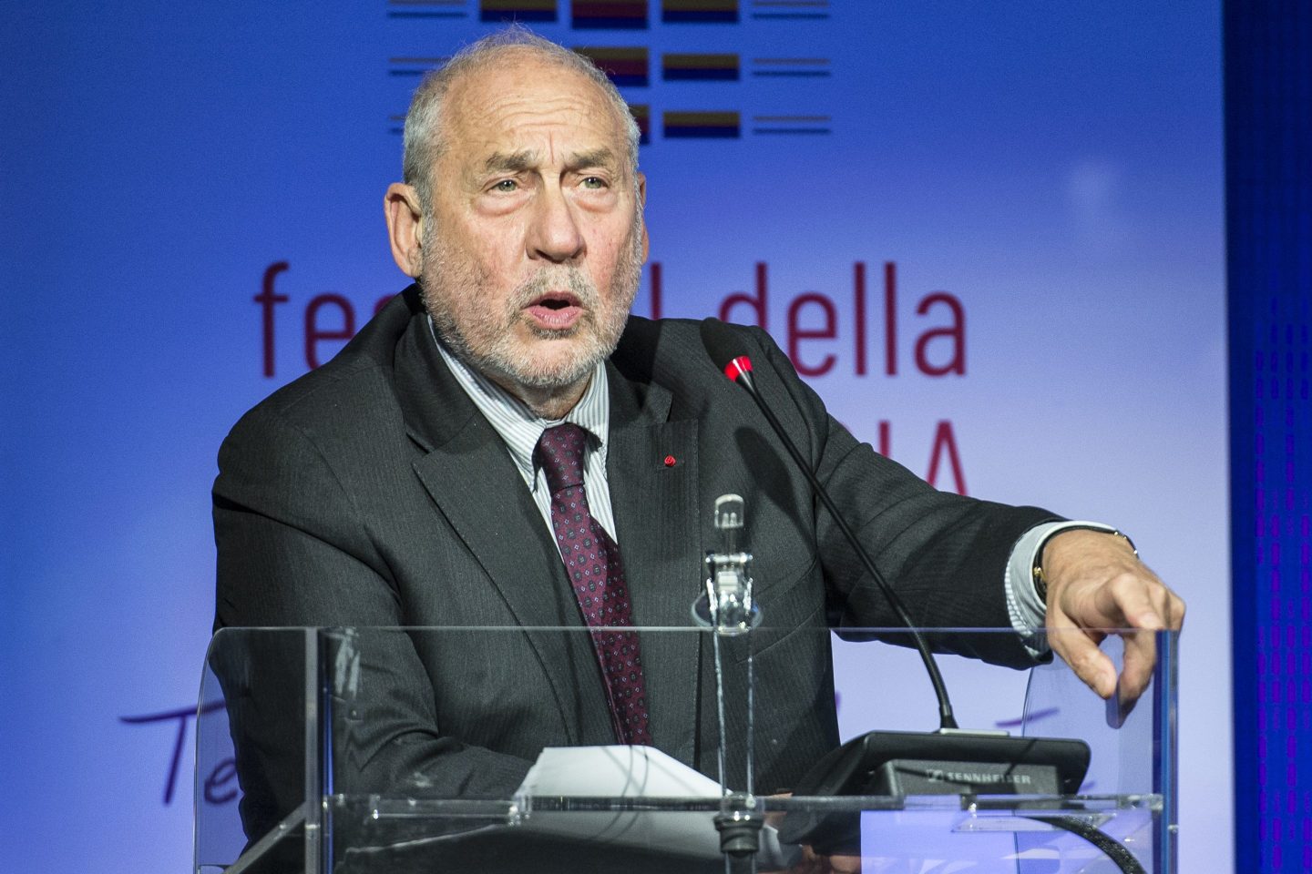 Joseph Stiglitz stands behind a glass podium, pointing emphatically.