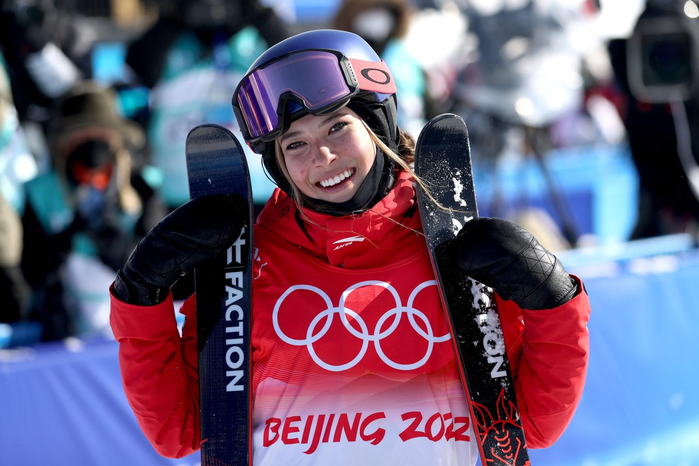 Eileen GU, wearing a red and while Beijing Olympics coat, smiles with her skis.