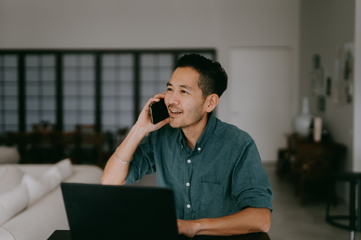 A man sits in his bedroom at a desk, looking at his laptop and taking a phone call.