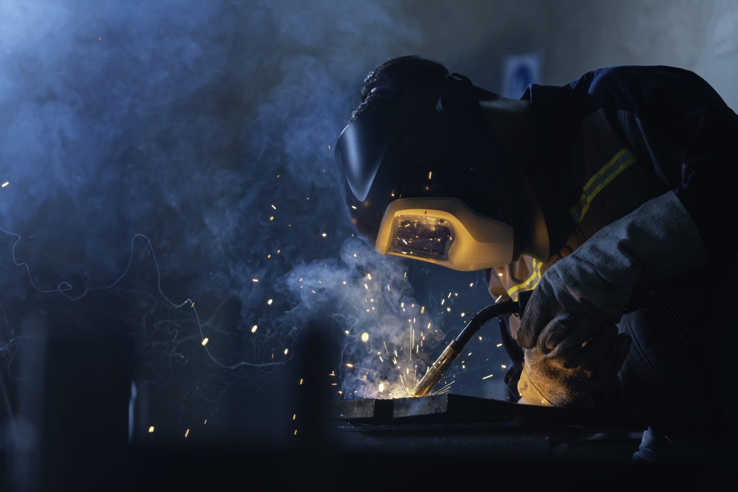 A professional welder wearing a dark helmet and gloves welds steel in a shadowy industrial setting, unleashing a cascade of bright, flying sparks.