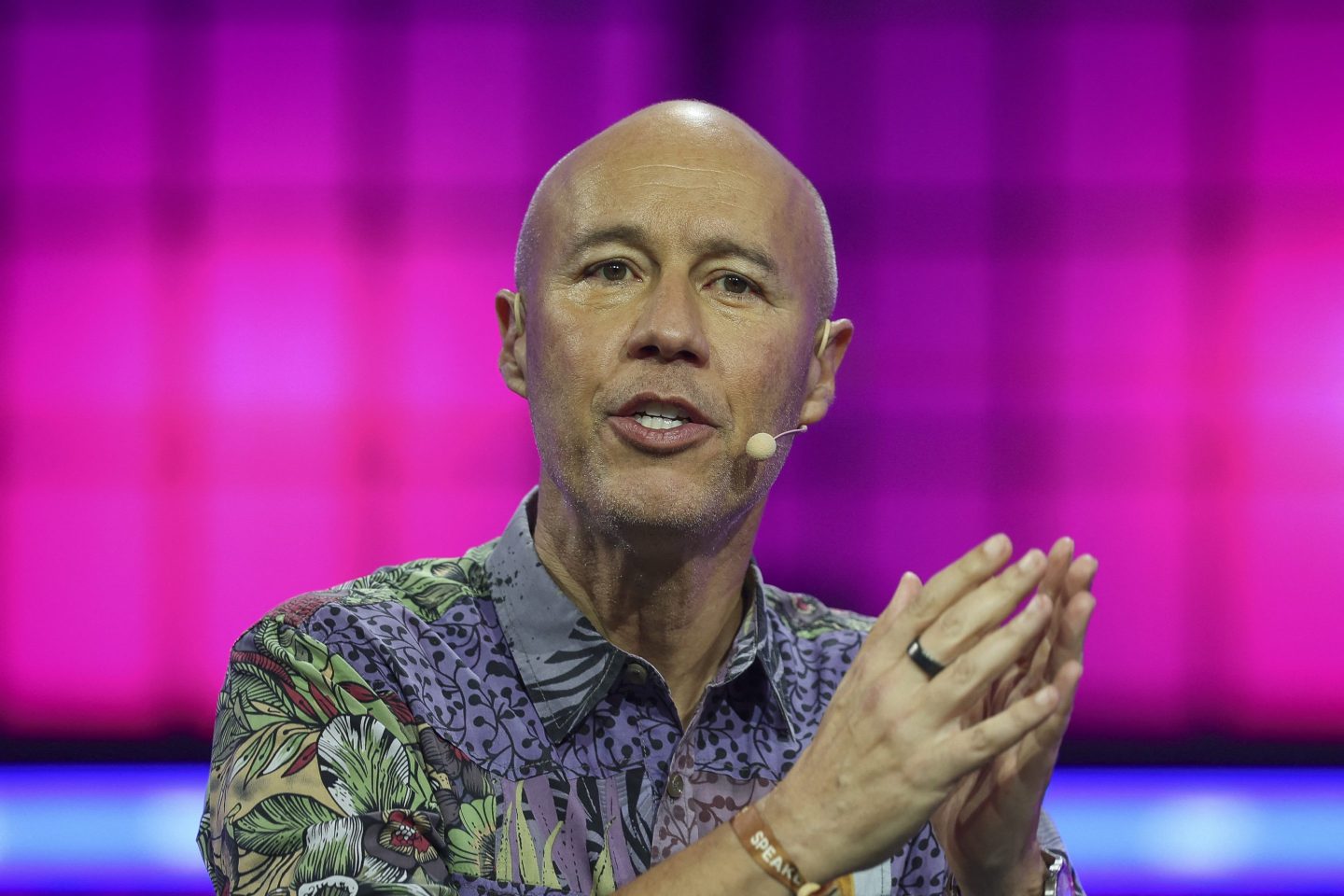 David Risher, wearing a patterned shirt, speaks in front of a bright magenta background.