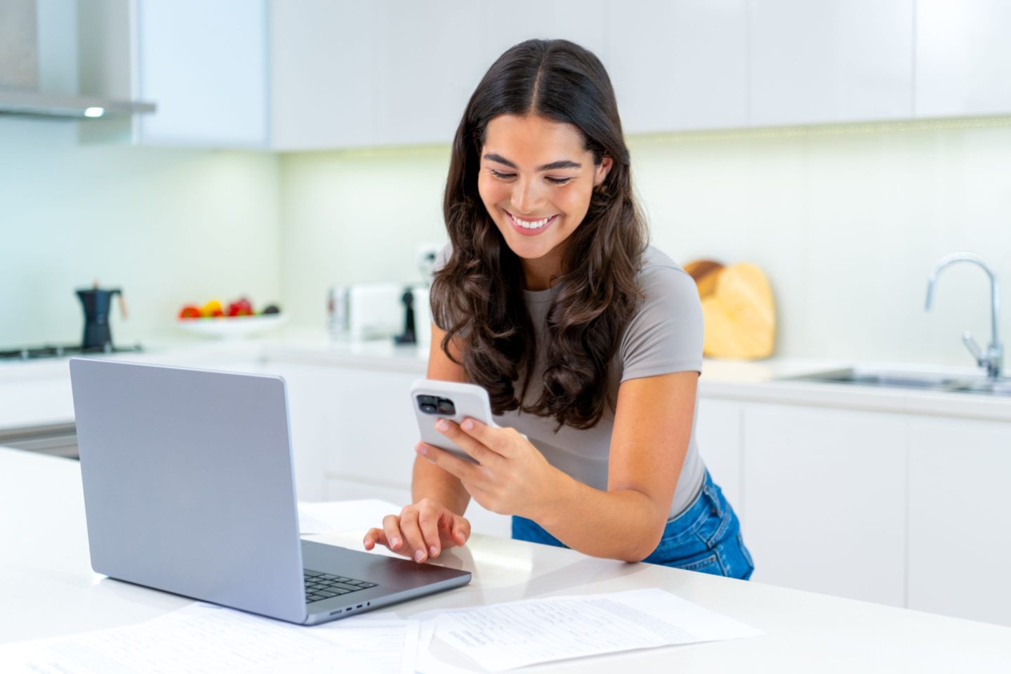A person looking at their phone and computer in a kitchen.
