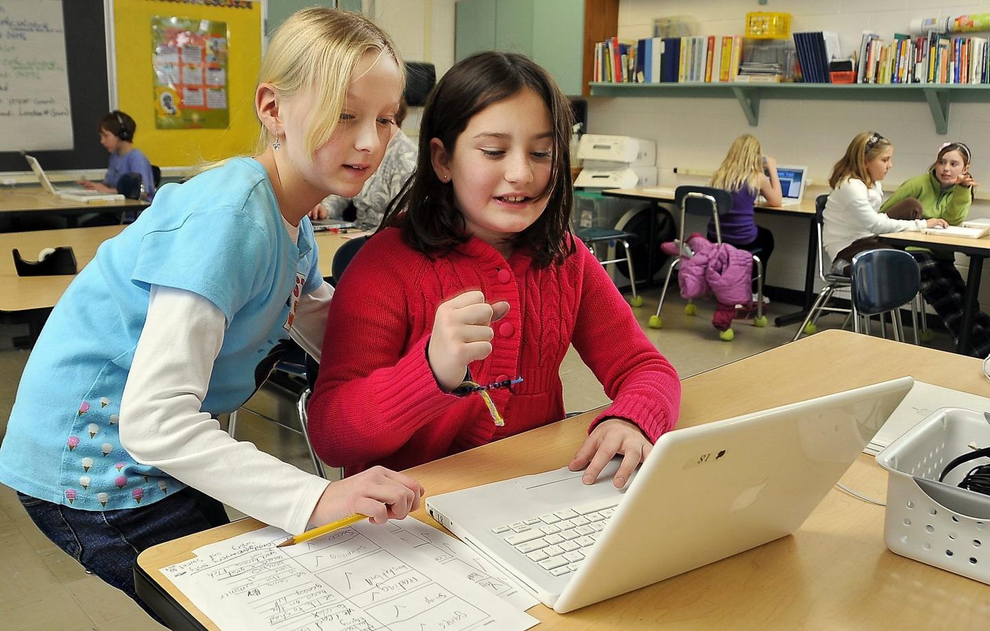 Two girls look at a laptop placed on a desk.