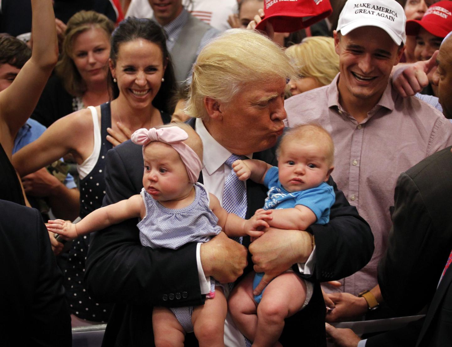 Donald Trump, holding two babies in his arms, leans over to kiss on on the head.