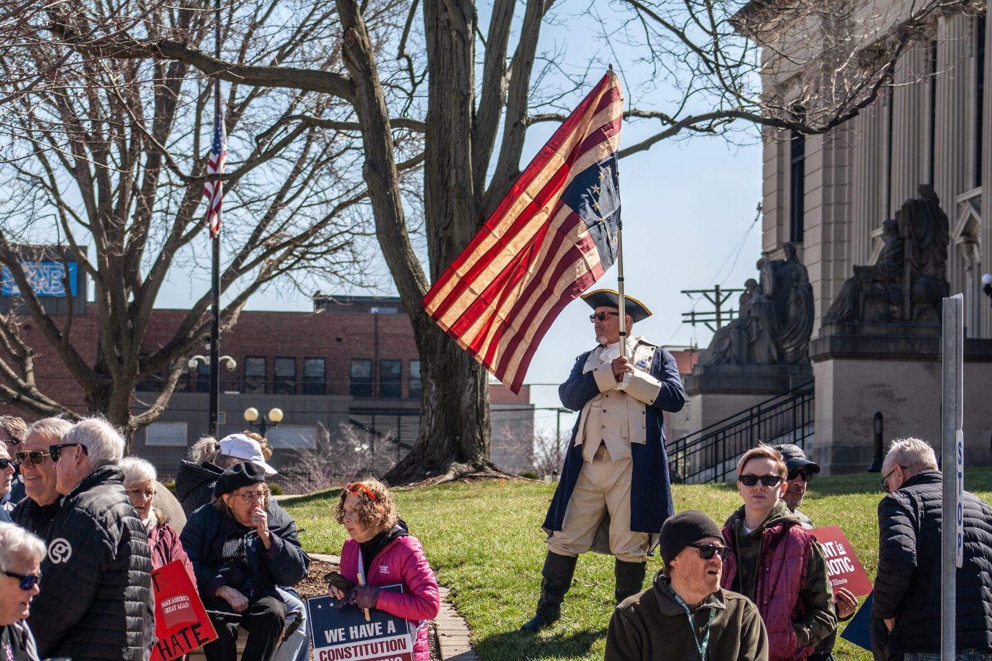 A demonstrator wearing a Revolutionary War uniform holds an upside-down Betsy Ross U.S. flag during a "No Kings" protest outside the Illinois State Capitol in Springfield, Ill., on Saturday, March 28, 2026. (Jenna Schweikert, Capitol News Illinois via AP)