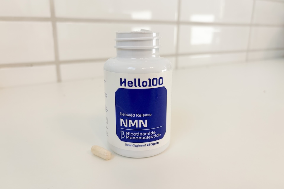 A bottle of Hello100 Delayed Release NMN sitting on a white countertop. One of the capsules is out of the bottle and placed next to it.