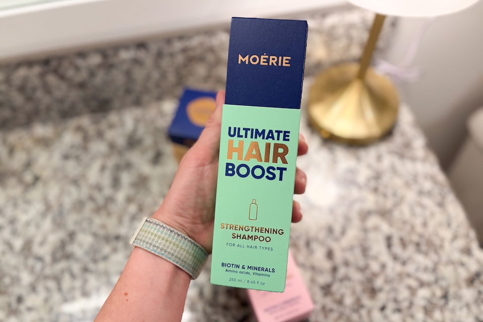 A close-up of a hand holding a bottle of Moerie Ultimate Hair Boost Strengthening Shampoo.
