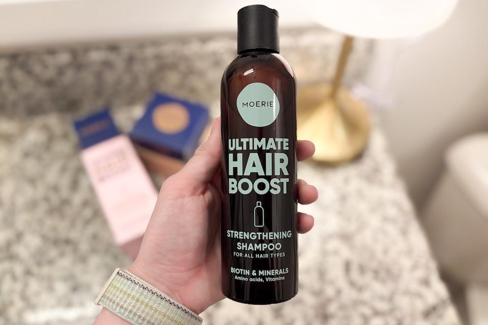 A close-up of a hand holding a bottle of Moerie Ultimate Hair Boost Strengthening Shampoo.