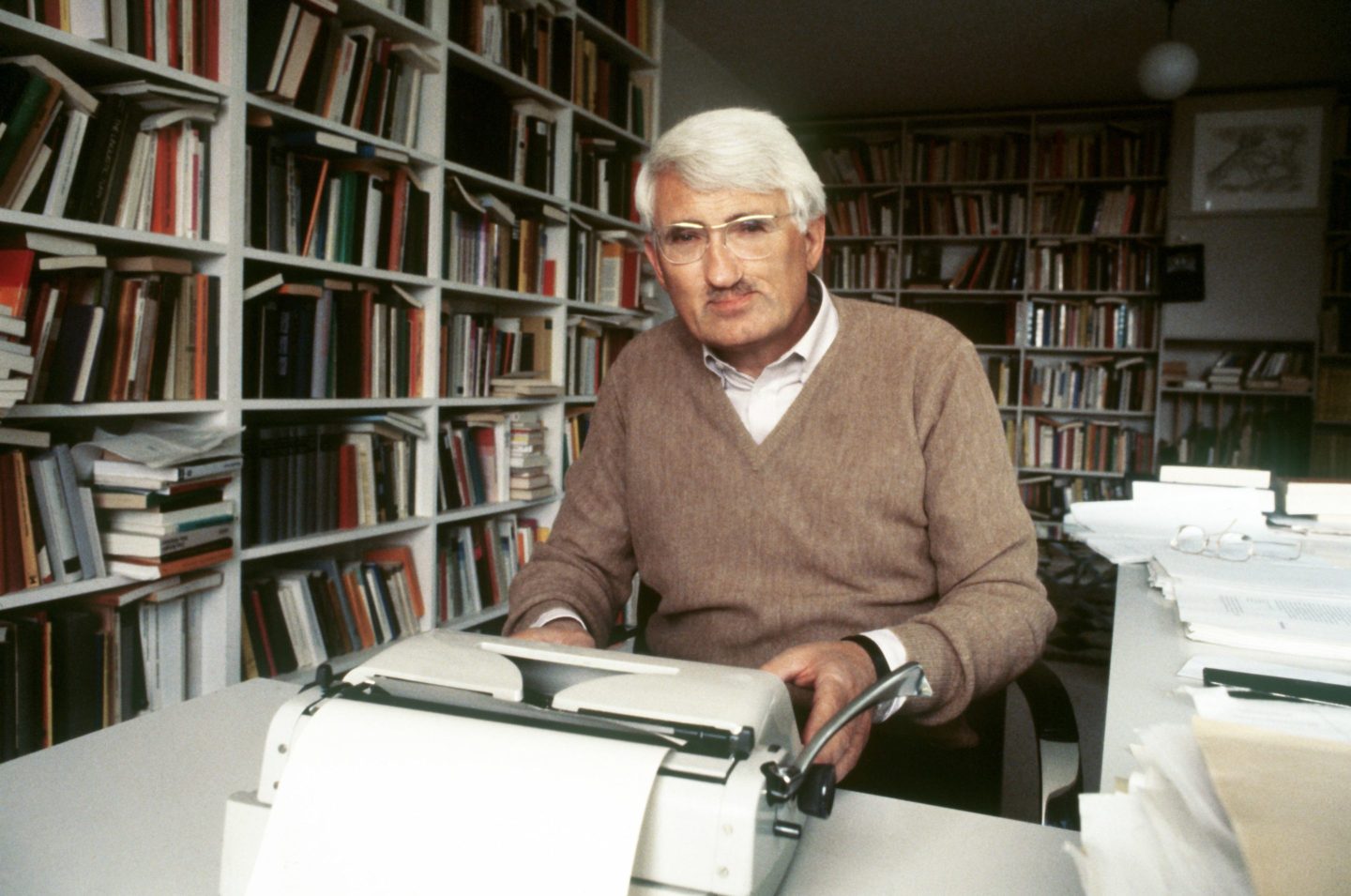 Habermas sits at a desk and types on a typewriter. He is in a room with full bookshelves on the walls