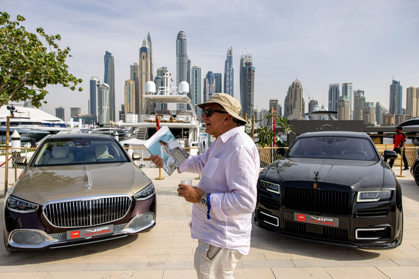 A man walks between two luxury cars with the skyline of Dubai in the background.