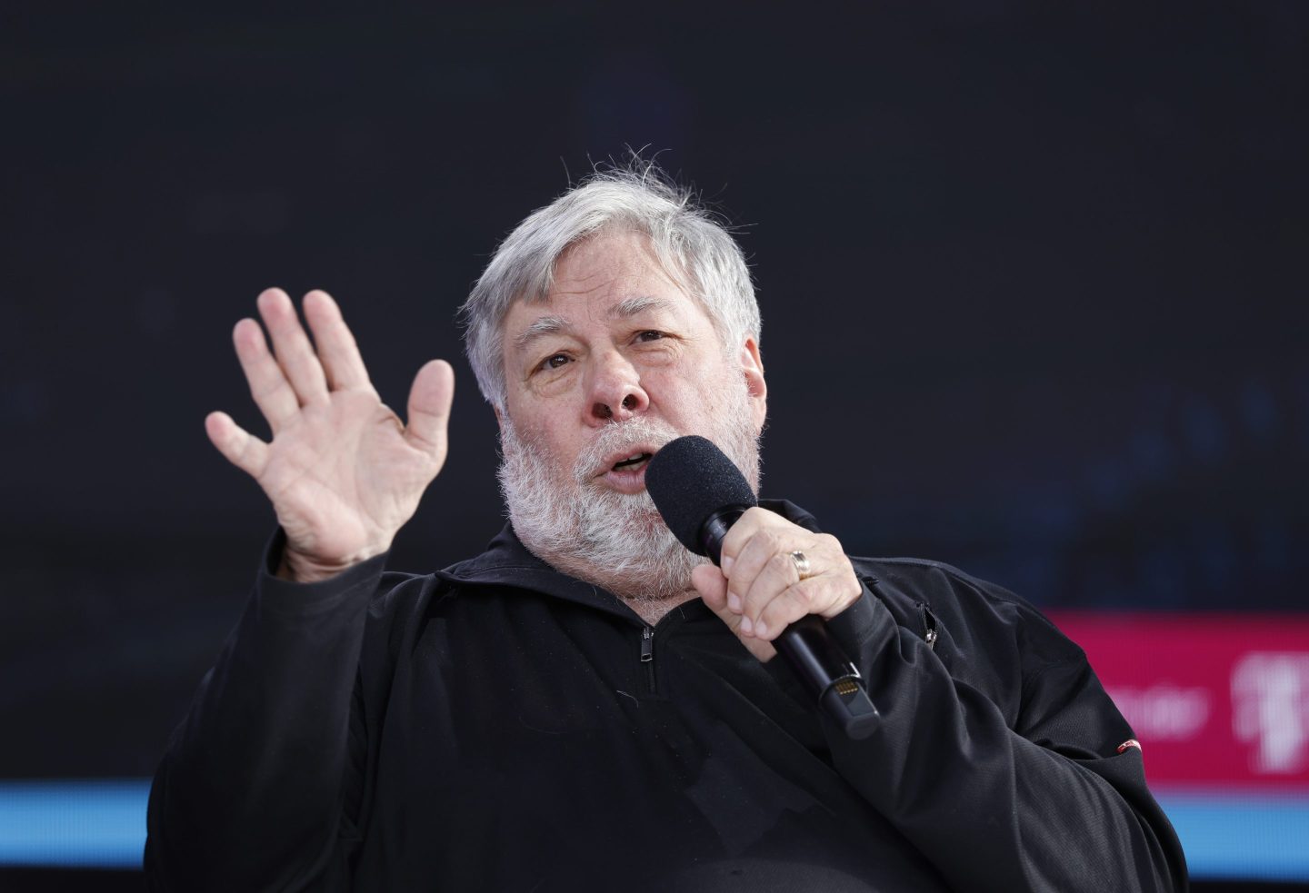 Steve Wozniak speaks into a microphone, raising his palm in the air.