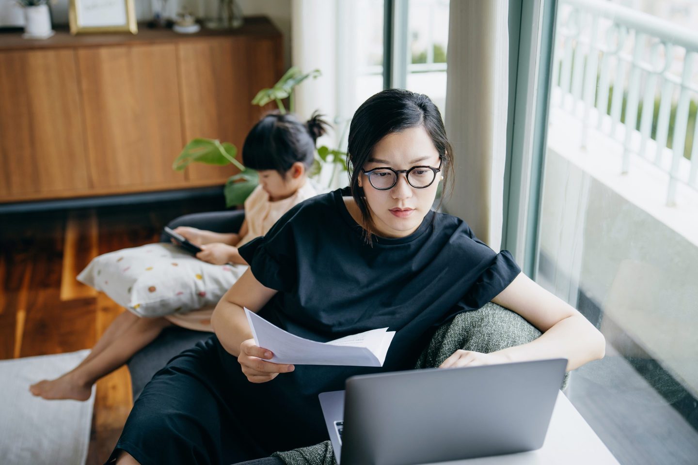 A mother works on her computer on a couch with her child in the background.