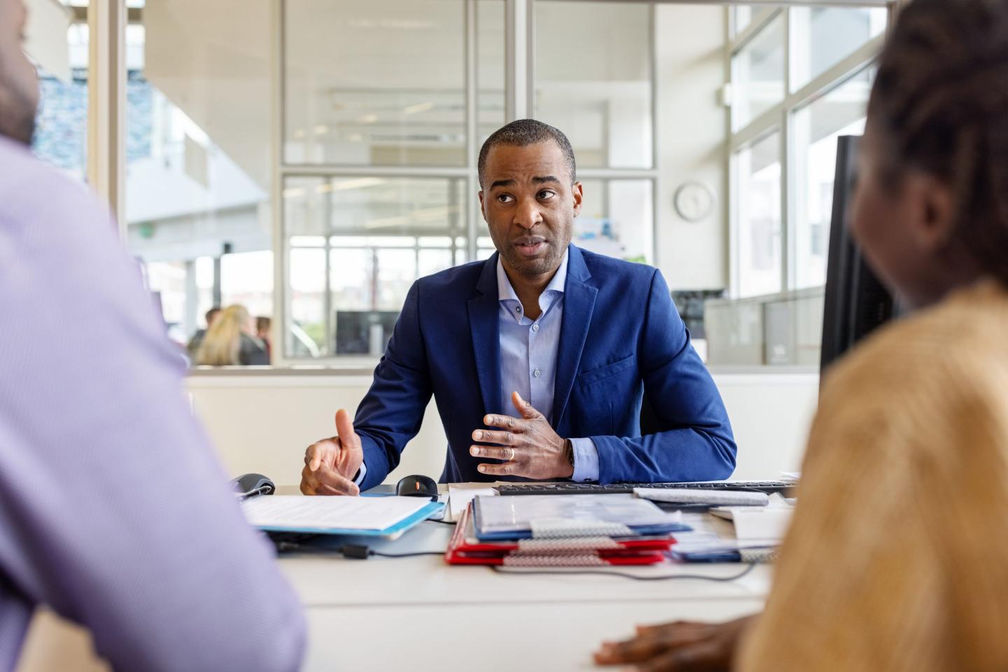 Picture of a man in a blue suit jacket and blue shirt sitting at a table consulting with two people partly visible in the foreground.