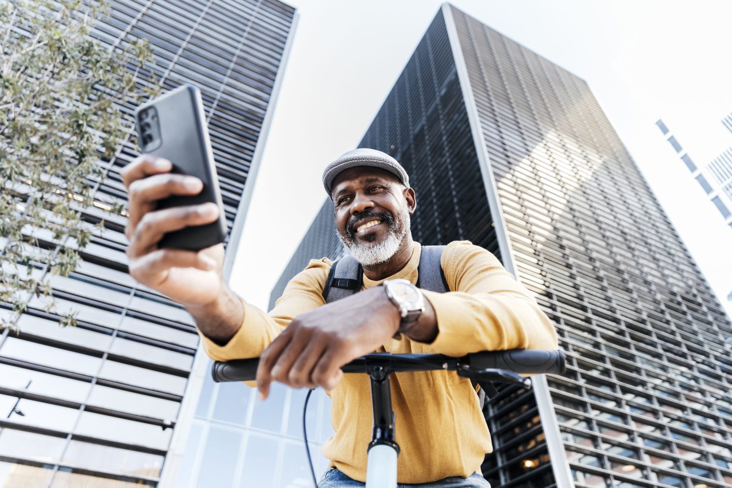 A man in his 50s is looking at his phone while standing on his bike in front of a modern skyscraper in the city