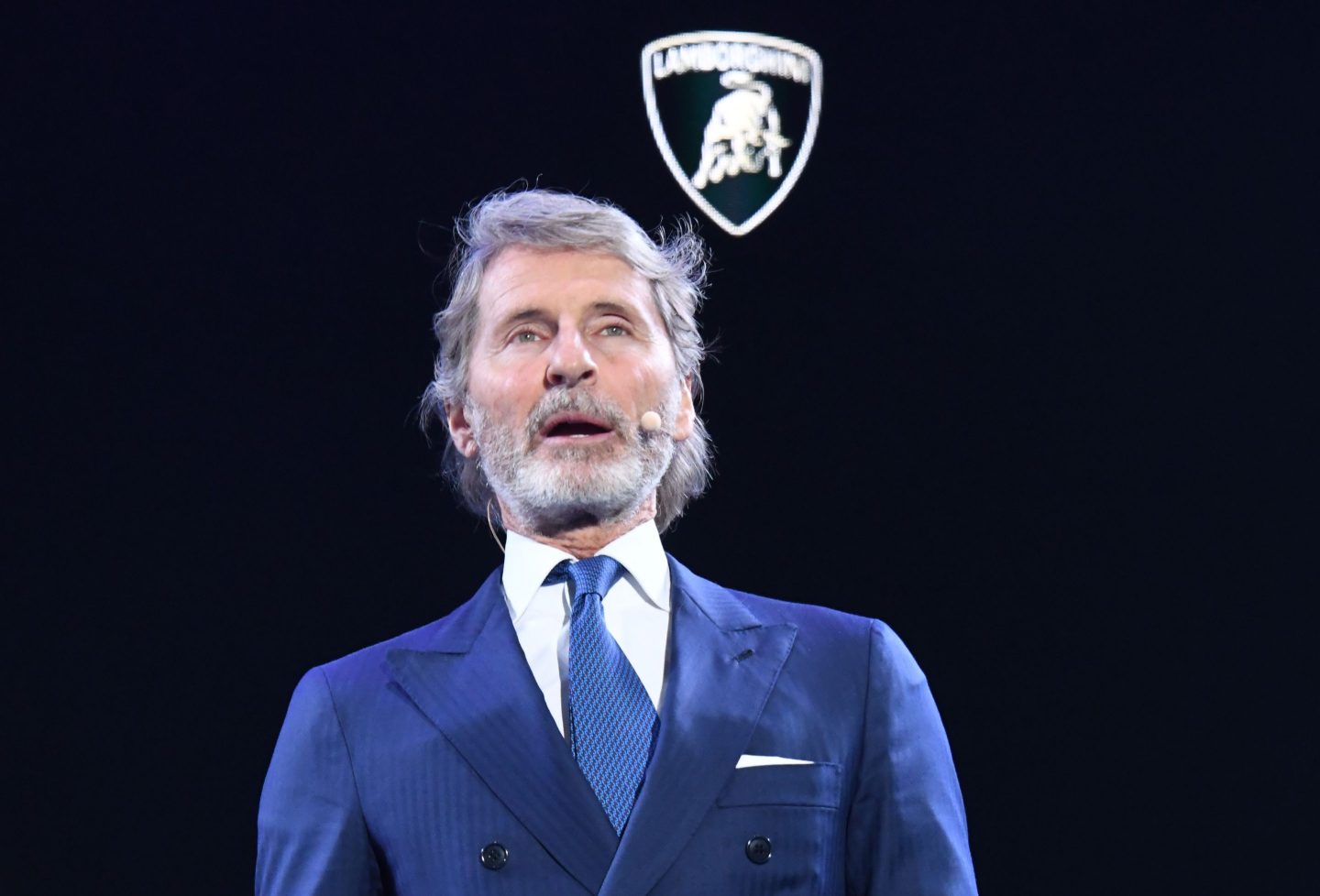 Stephan Winkelmann, wearing a dark blue suit, speaks in front of a black background with the Lamborghini logo on it.