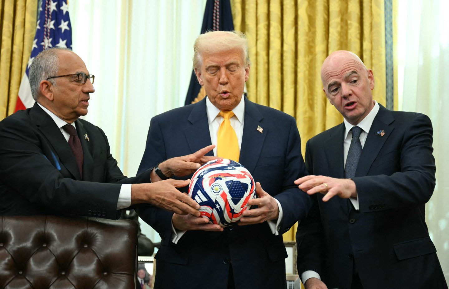 Trump holds a soccer ball along with two men in suits on either side of him.