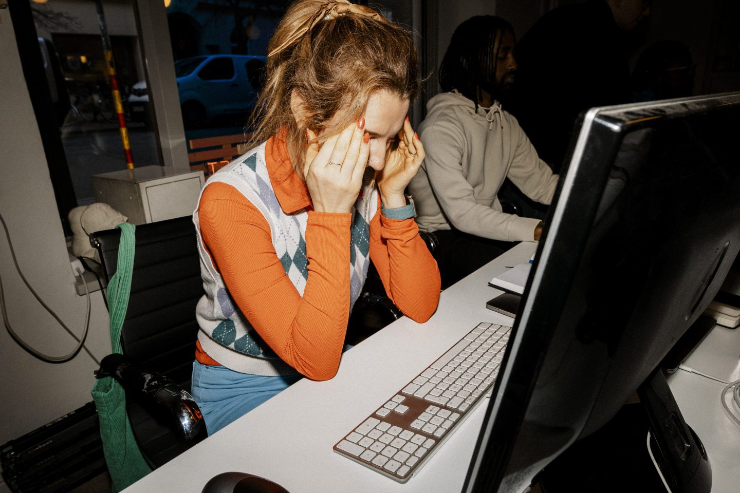 A woman in an orange shirt sits by a computer with her fingers rubbing her temples.