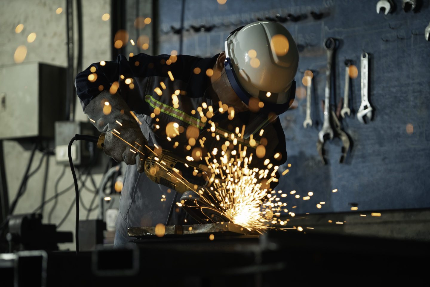 A skilled artisan wearing helmet and goggles grinds a steel surface, creating lively trails of sparks in a low-lit manufacturing environment.