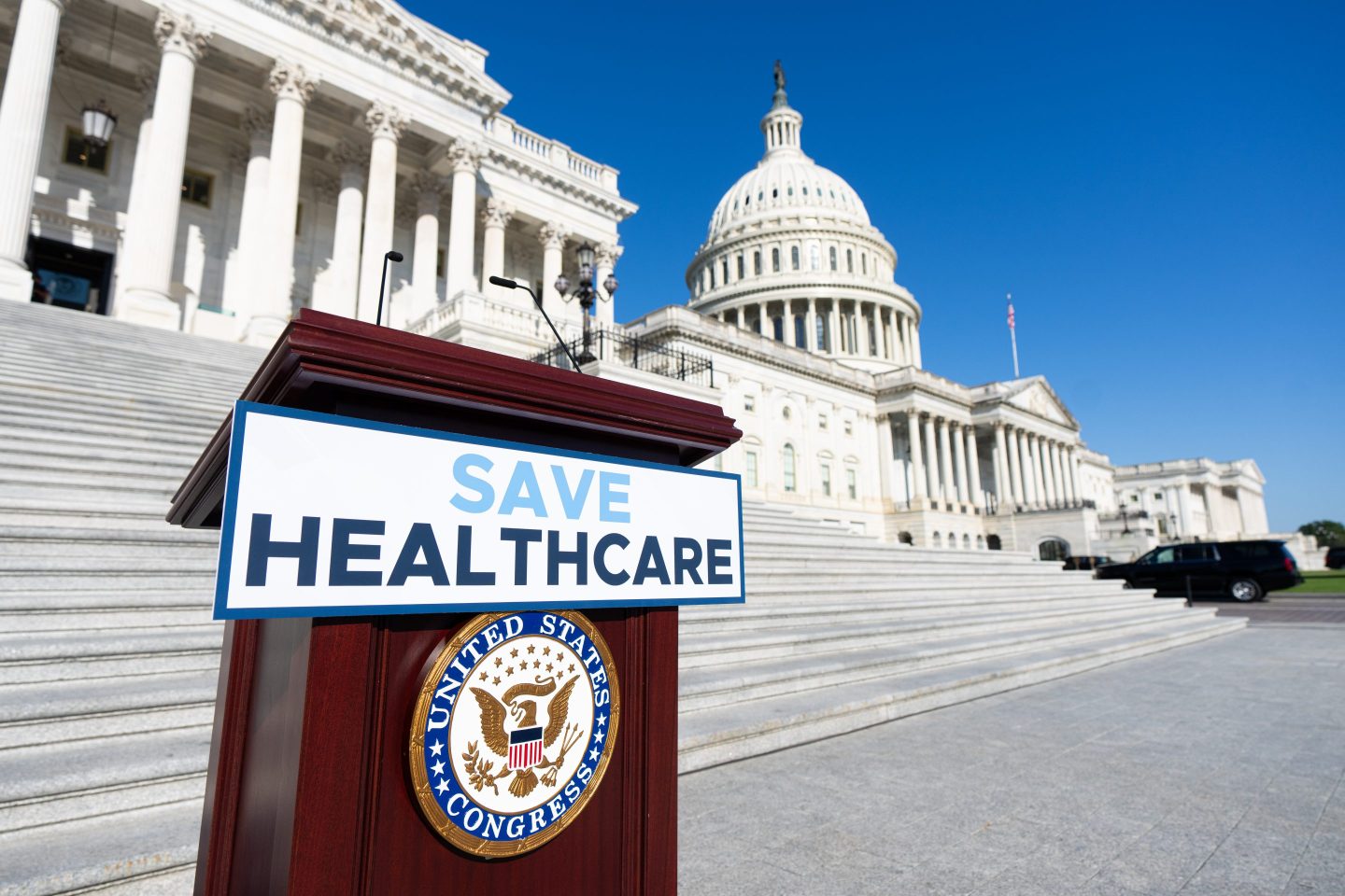 A podium with a sign reading "save healthcare" stands in front of the U.S. Capitol building.