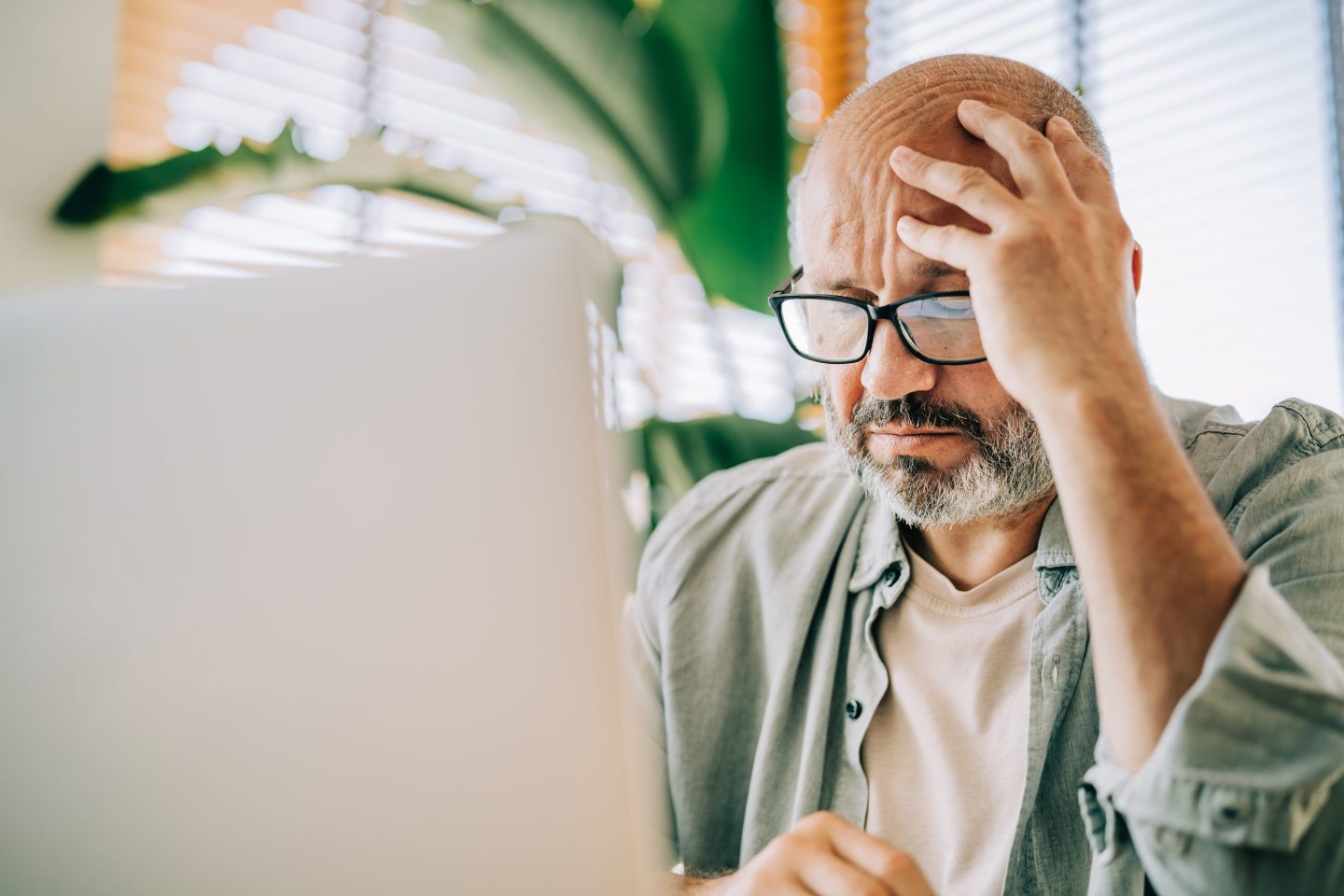 A close-up portrait of a thoughtful middle-aged man wearing glasses, looking intently at his laptop screen.