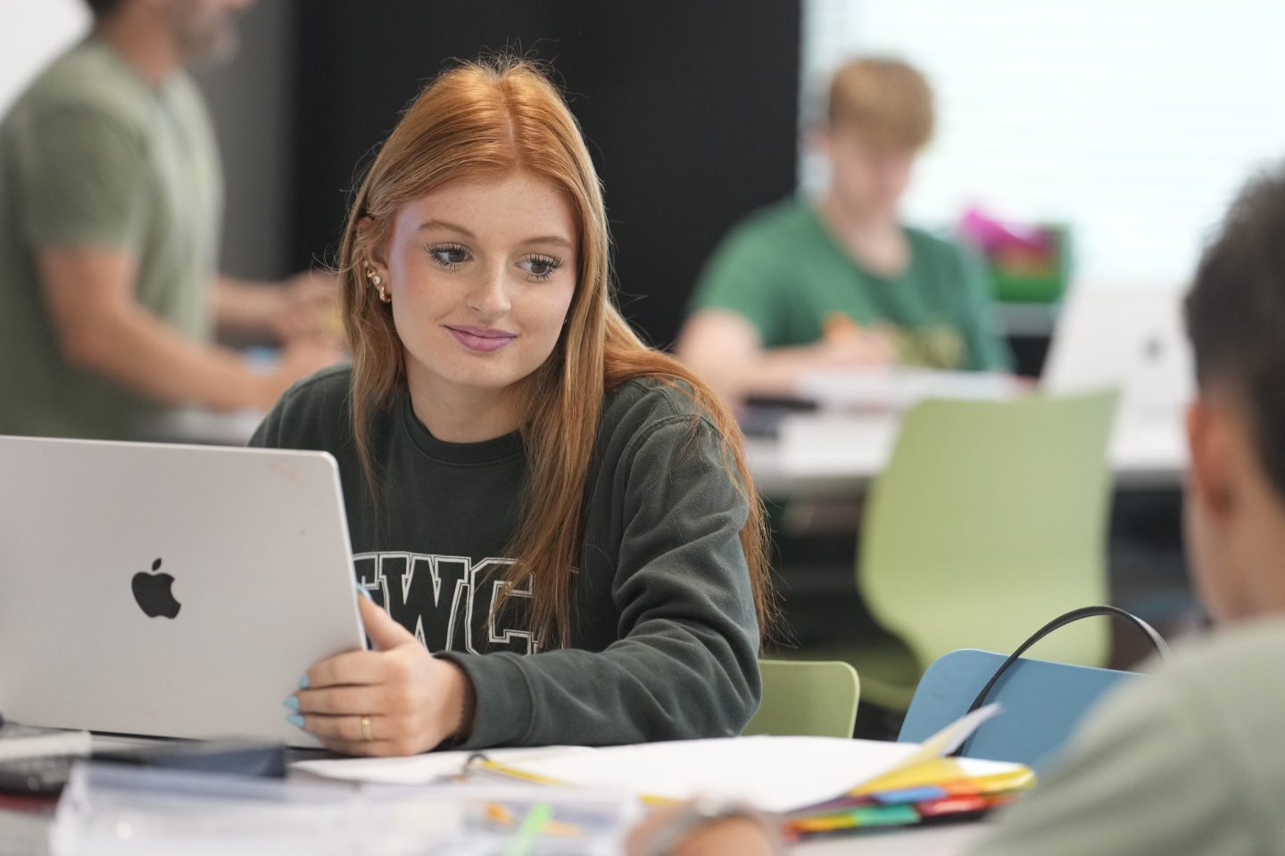 A girl with red hair sitting at a desk looks up from the laptop in front of her.