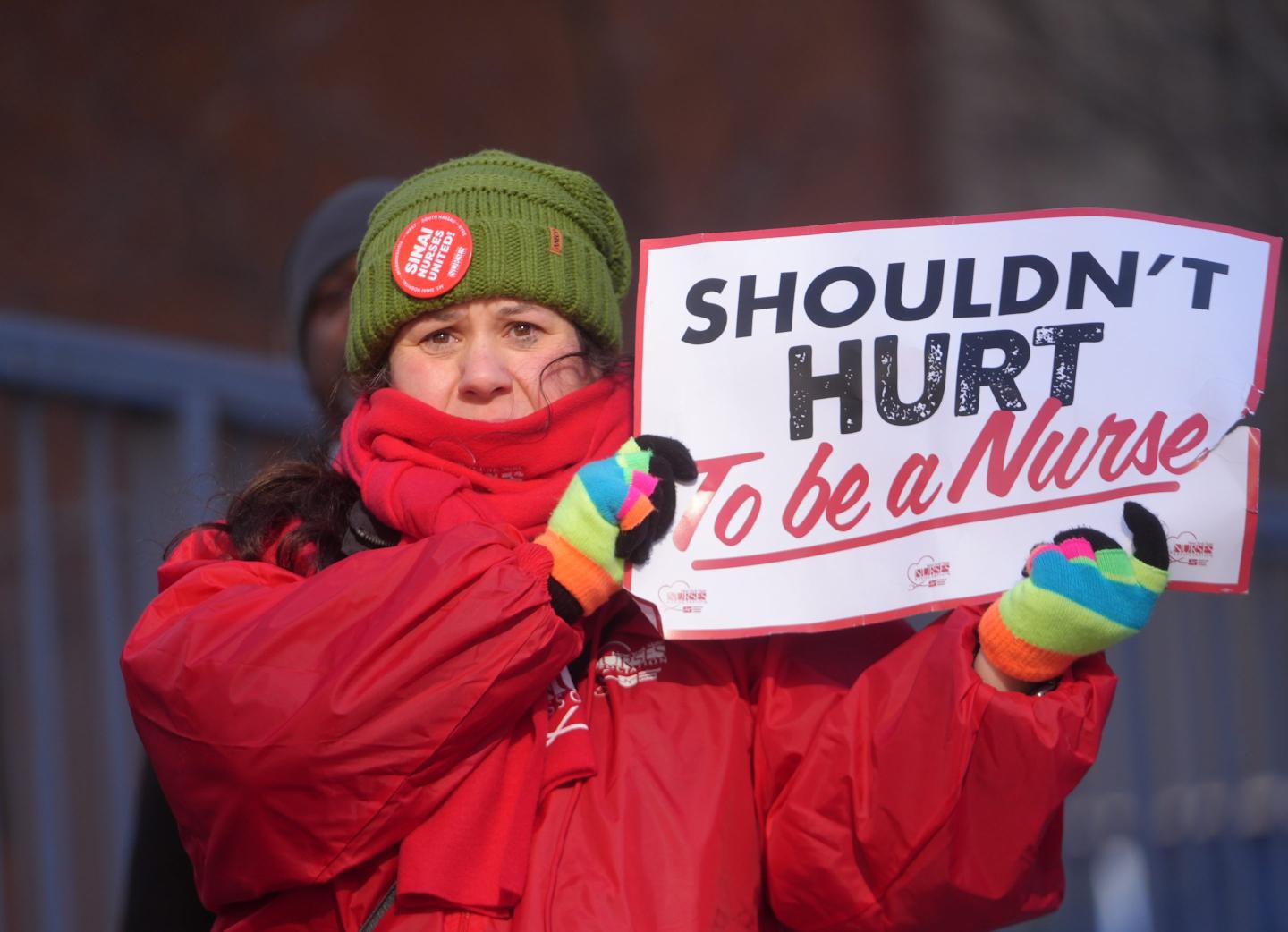A woman in a red coat holds up a sign that says, “Shouldn’t hurt to be a nurse.”