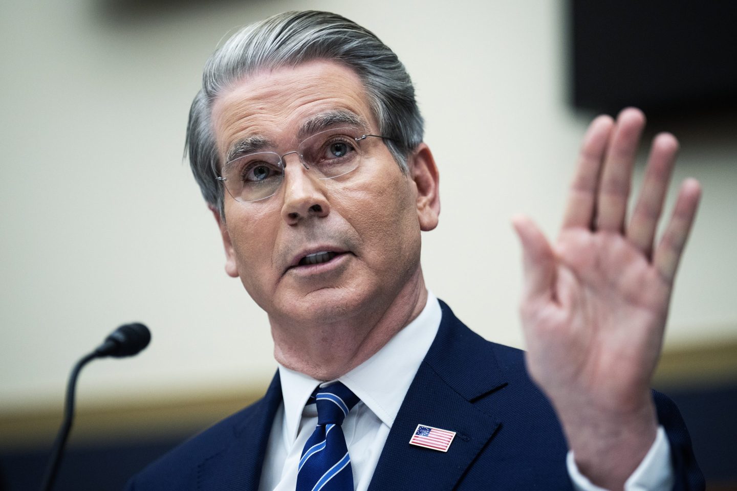 Scott Bessent, speaking into a microphone, raises one hand next to his face.