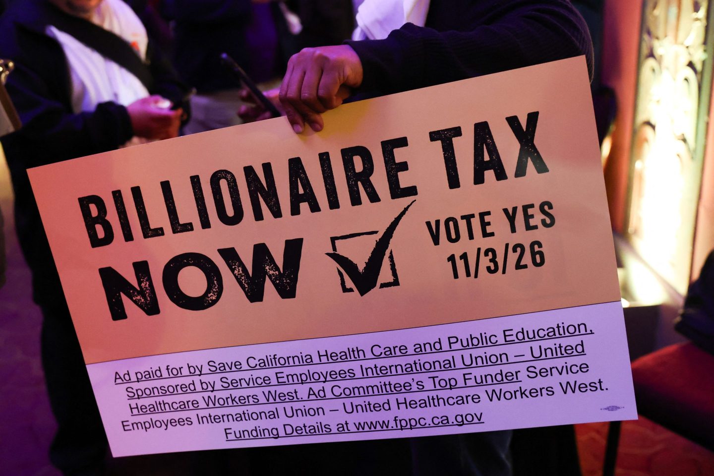 An attendee holds a sign reading "Billionaire Tax Now" in all caps.