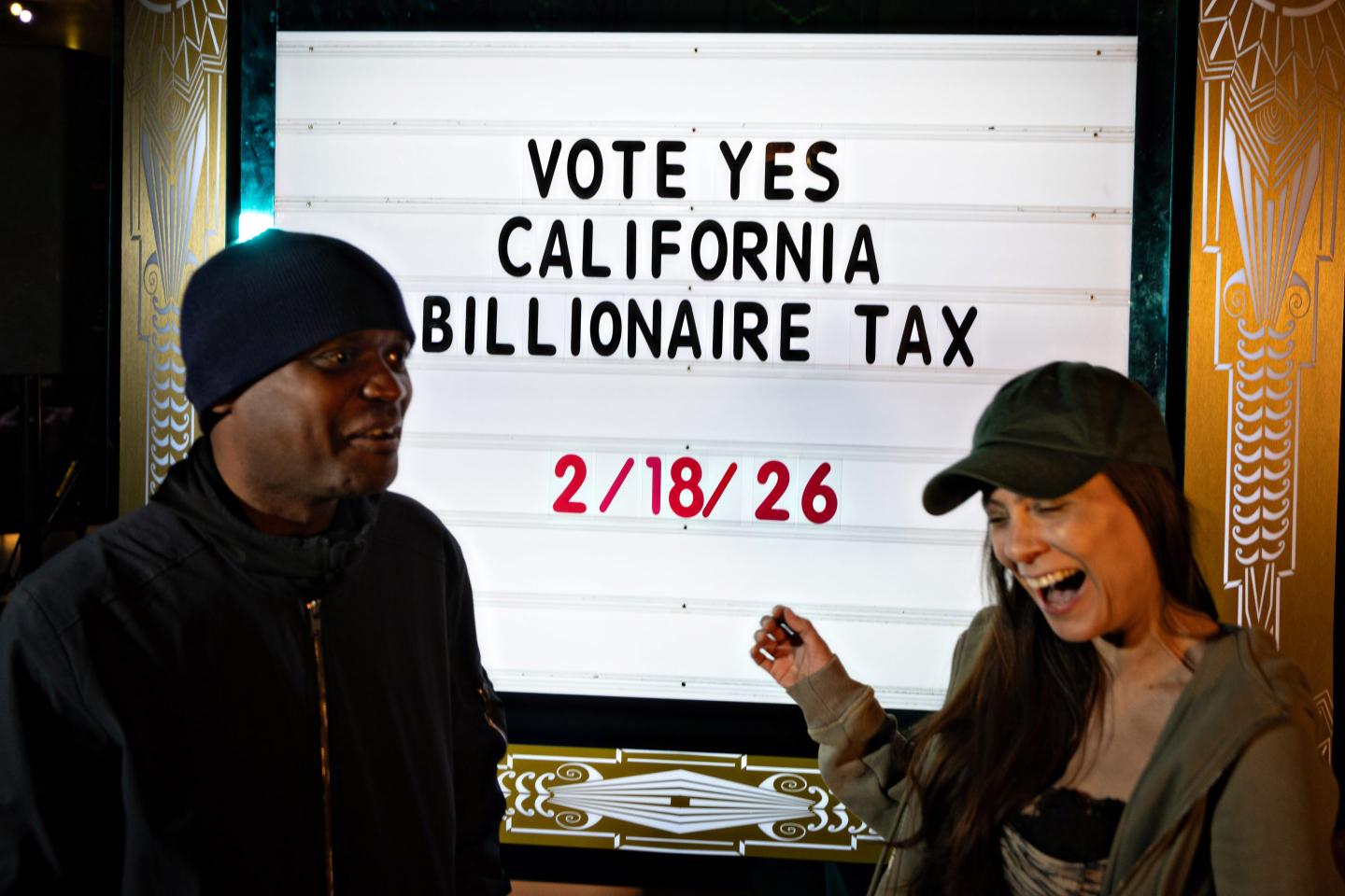 Two people stand in front of a sign urging support for California’s billionaire tax