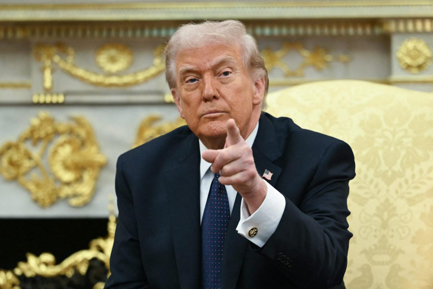 Donald Trump sits in the Oval Office. He looks ahead and points.