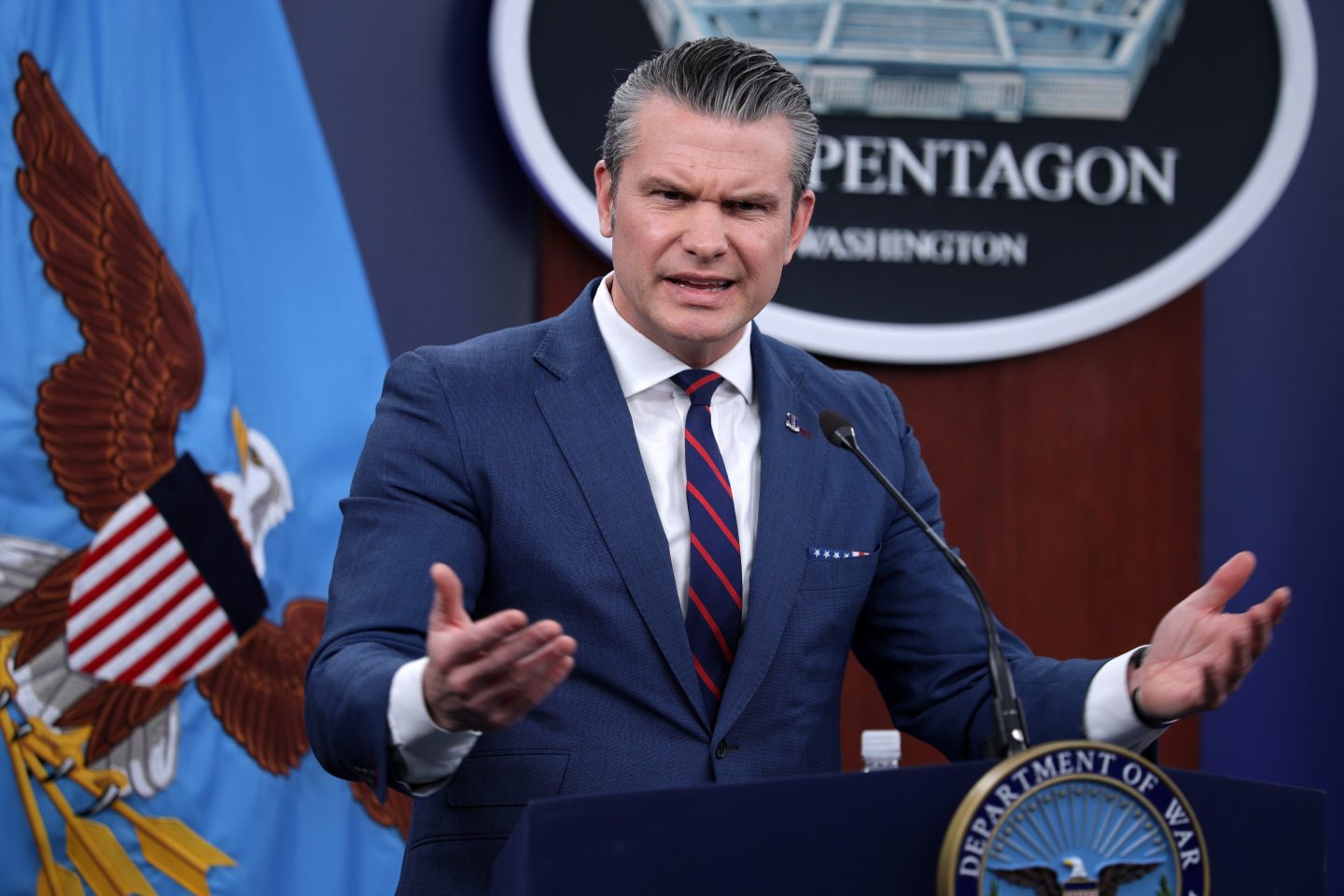 U.S. Secretary of War Pete Hegseth standing in front of a podium with a Pentagon sign behind him, gesturing with his hands outstretched and looking angry.