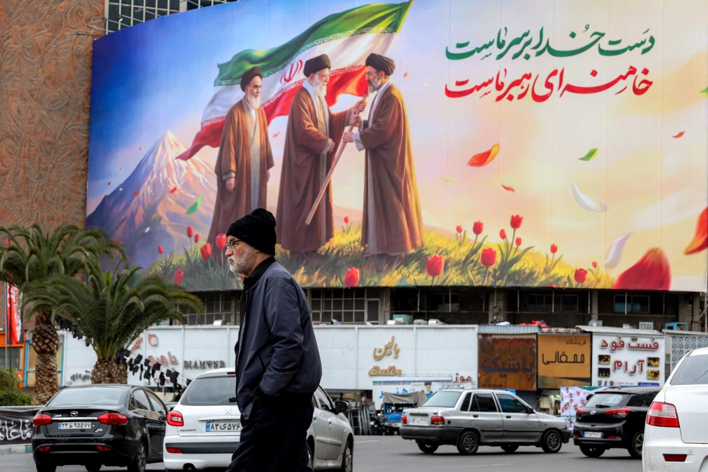 Photo: A man walks near a banner displayed at Valiasr Square in central Tehran on March 10, 2026, depicting Iran's late supreme leader Ayatollah Ruhollah Khomeini (L) watching as his successor the late Ayatollah Ali Khamenei (C) hands over a national flag to his son and new supreme leader Mojtaba Khamenei (R). Iran marked the appointment of Ayatollah Mojtaba Khamenei to replace his father as its supreme leader with a new barrage of missiles against Israel and the Gulf states on March 9, as the Middle East war sent oil prices soaring. (Photo by AFP) / Attention editors: AFP covers the war in the Middle East through its extensive regional network, including bureaus in Tehran, Jerusalem, and several neighboring countries. Since the start of the conflict, journalists have been working under increasingly restrictive conditions. Authorities in several countries have limited reporters' movements, photo and live video coverage from sensitive locations. Some governments and armed groups have banned images of missile or drone strikes and other security-related sites. /