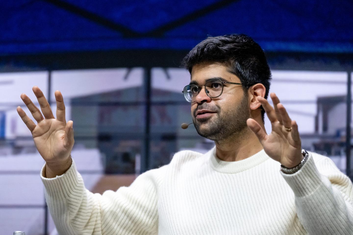 Aravind Srinivas, wearing a white sweater, lifts both of his arms in front of him.