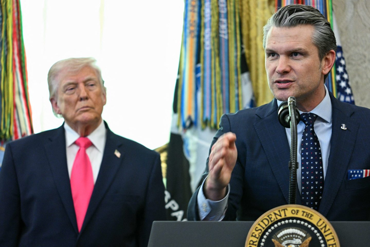 Pete Hegseth speaks behind a podium as Donald Trump watches behind him.