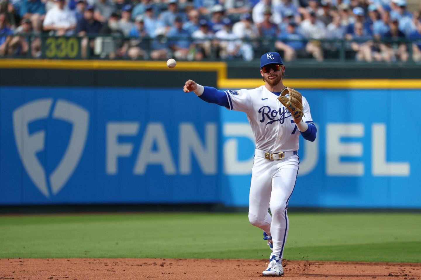 Bobby Witt Jr. throws a baseball. He is standing in front of a FanDuel sign on a baseball field.