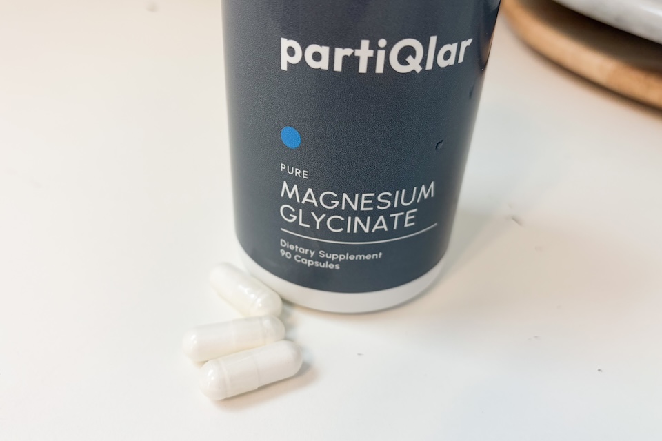 Three off-white capsules of partiQlar Magnesium Glycinate resting next to the bottle on a white countertop.