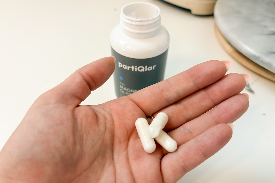 An open palm holding three off-white capsules of partiQlar Magnesium Glycinate.