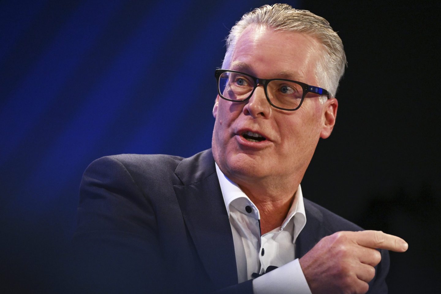 Ed Bastion, wearing a suit and glasses, speak and points with one finger to his left.