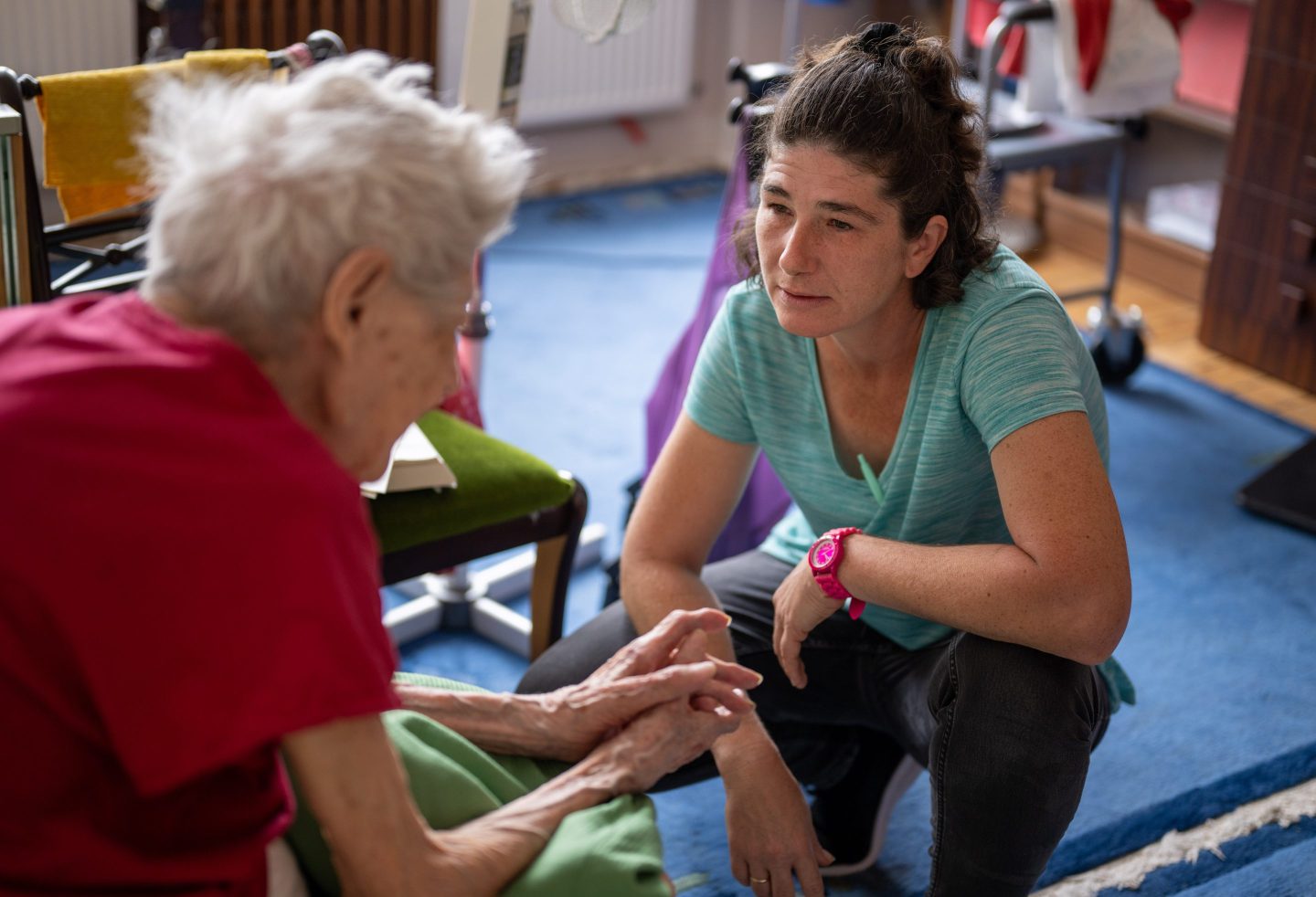 A woman kneels on the floor next to an older woman sitting down.